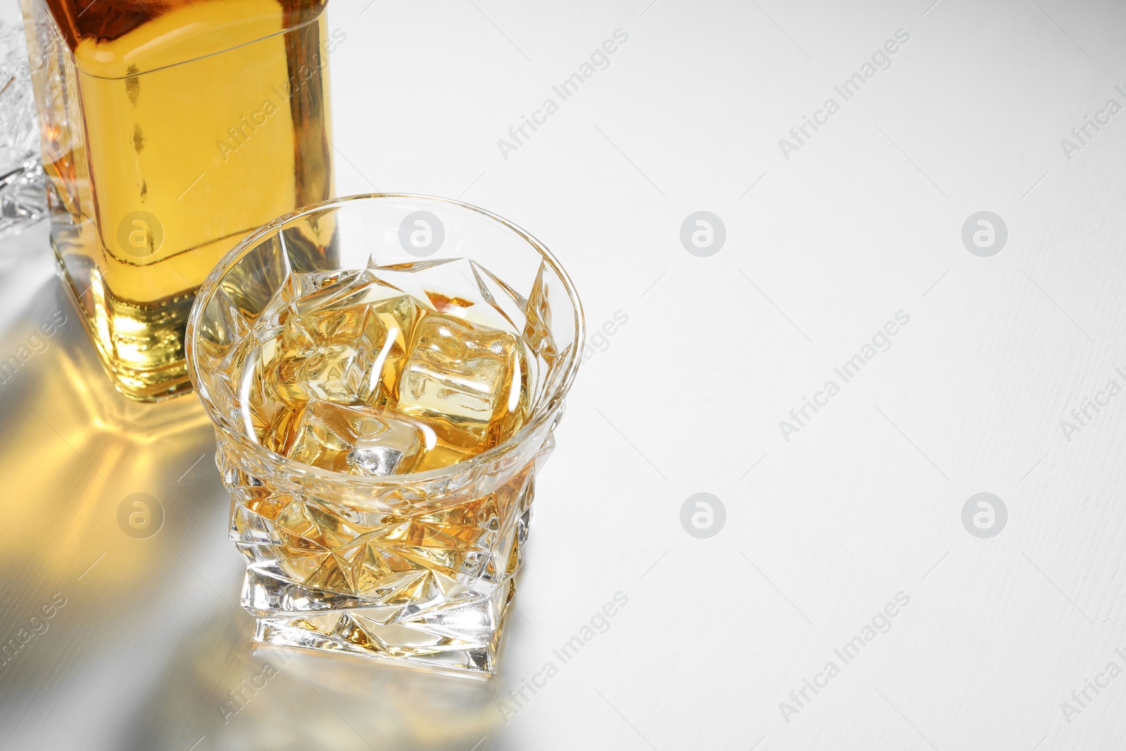 Photo of Whiskey with ice cubes in glass and bottle on white table, space for text