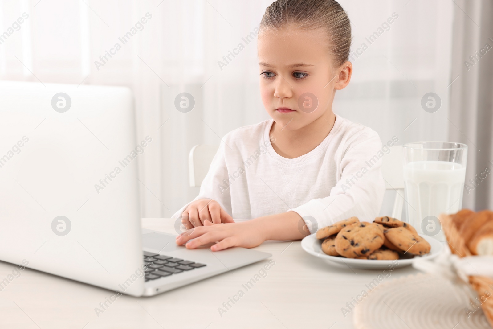 Photo of Little girl using laptop while having breakfast at table indoors. Internet addiction