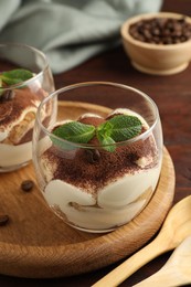 Delicious tiramisu in glasses, mint leaves, coffee beans and spoons on wooden table, closeup