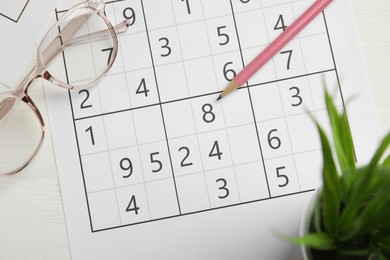 Photo of Sudoku, eyeglasses and pencil on white wooden table, flat lay