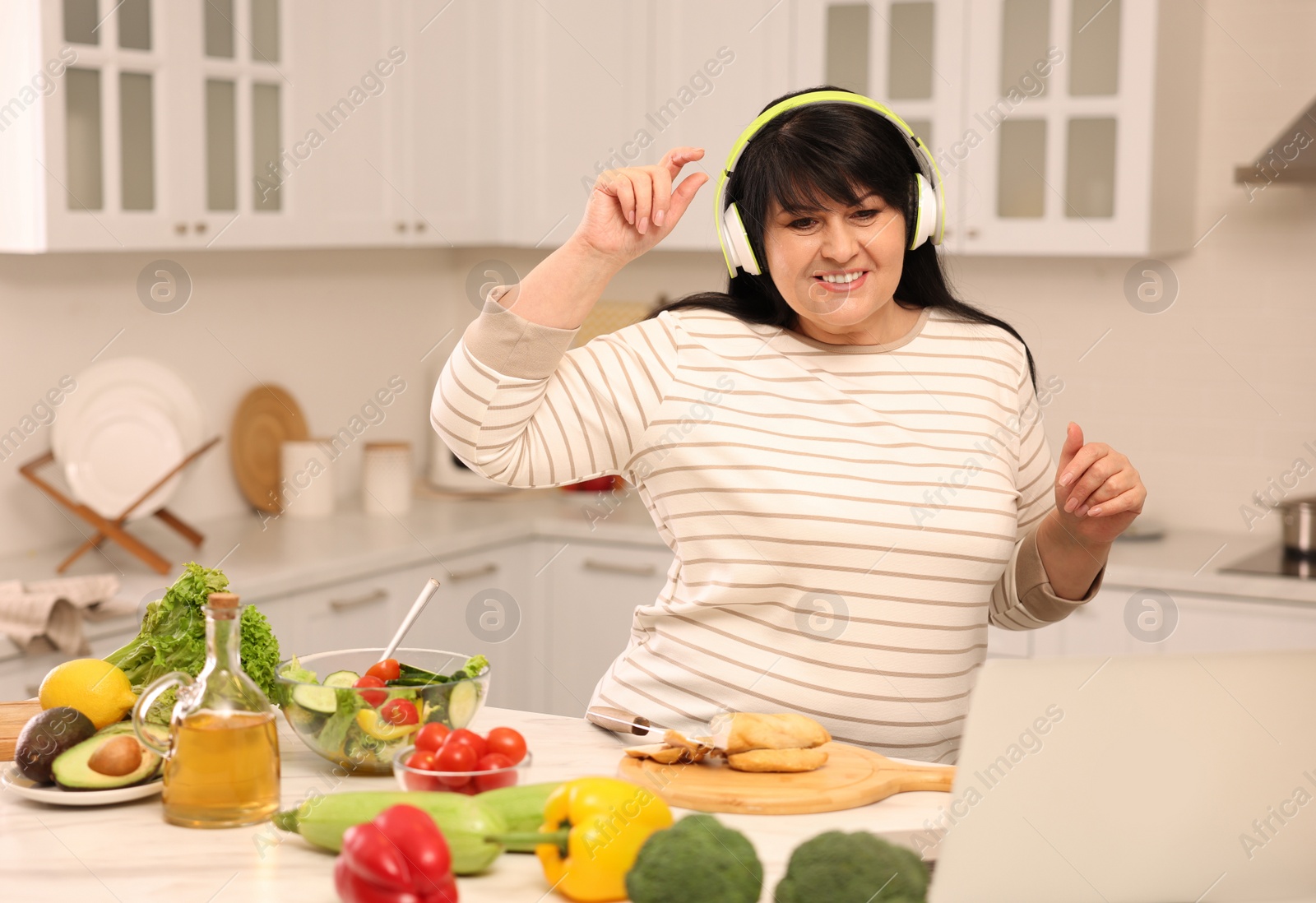 Photo of Happy overweight woman with headphones dancing while cooking in kitchen