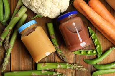 Jars with baby food and fresh vegetables on wooden table, flat lay