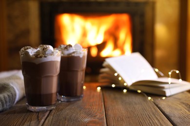 Photo of Glasses with hot cocoa, marshmallows, lights and book on wooden table near fireplace