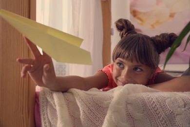 Cute little girl playing with paper plane on bed in room
