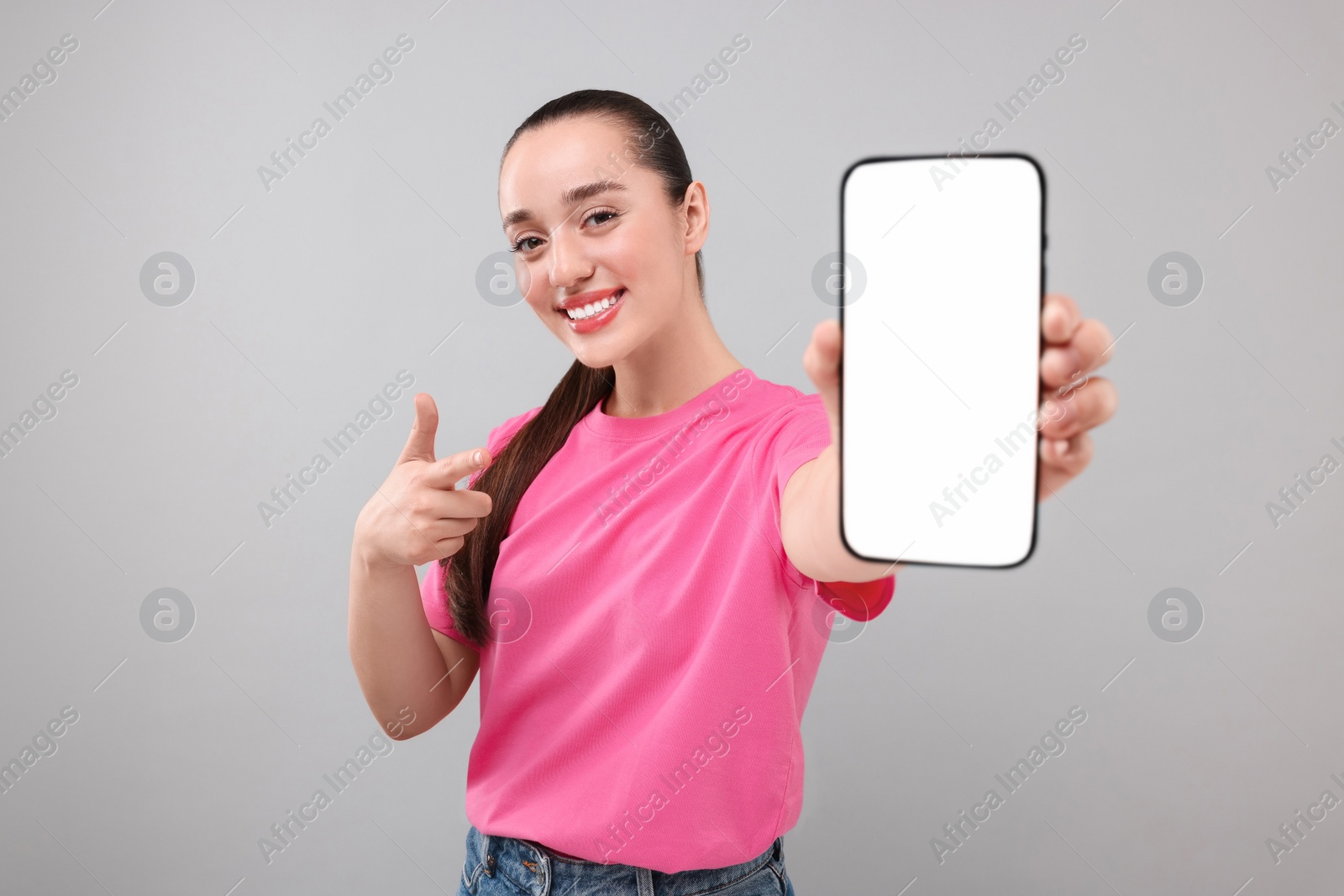 Photo of Young woman showing smartphone in hand and pointing at it on light grey background