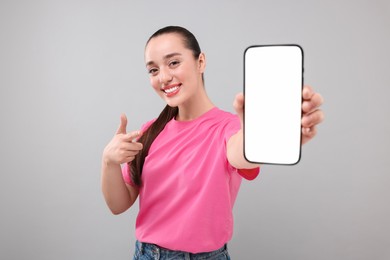 Photo of Young woman showing smartphone in hand and pointing at it on light grey background
