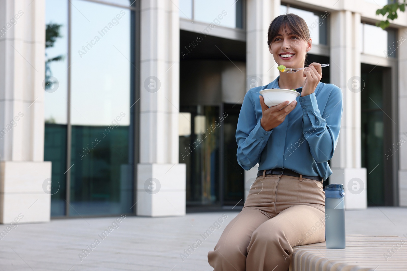 Photo of Happy businesswoman with plastic bowl of salad and bottle of water having lunch on bench outdoors