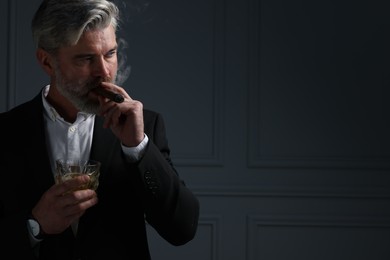 Photo of Handsome bearded man with glass of whiskey smoking cigar against dark grey background. Space for text