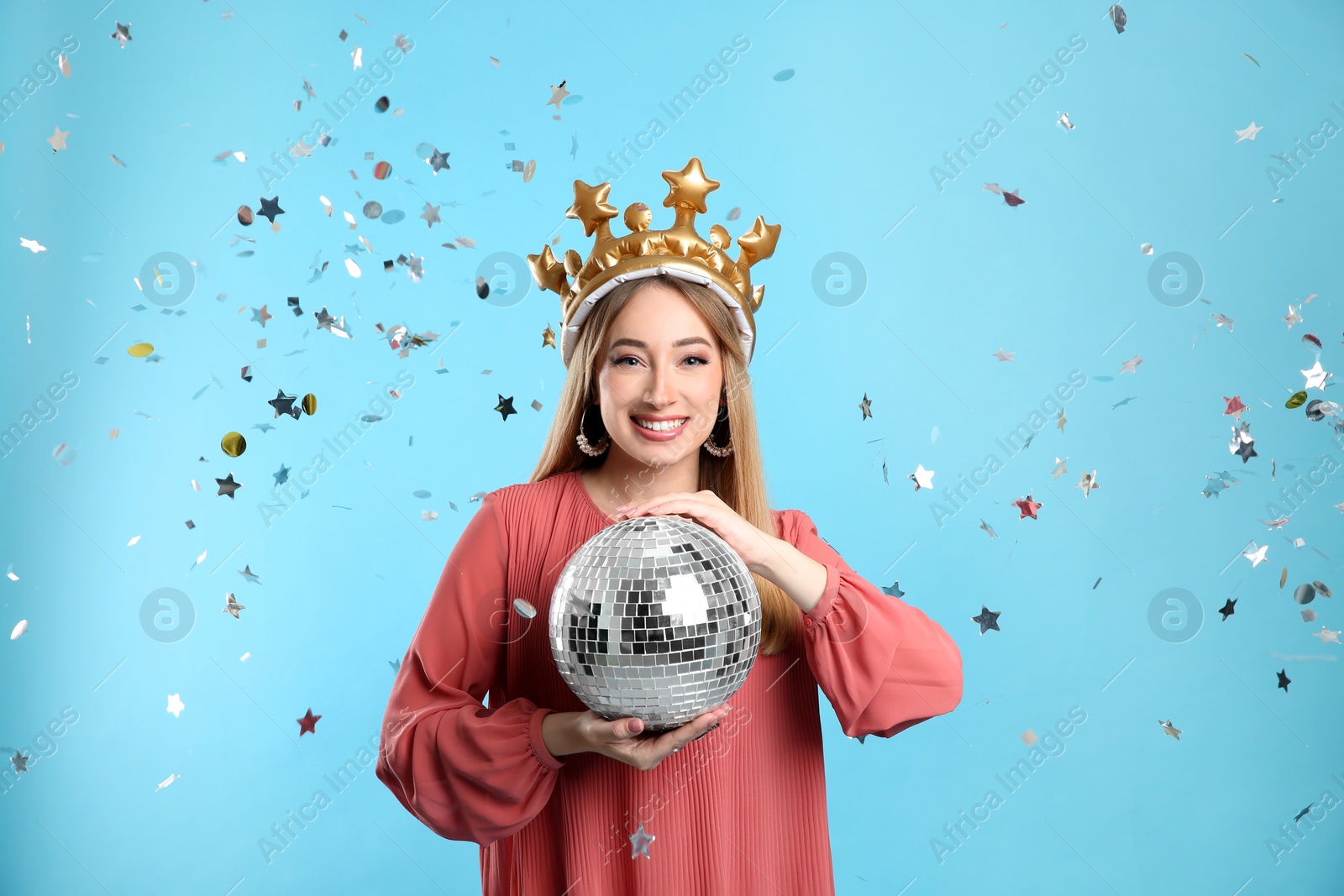 Photo of Happy young woman in party crown with disco ball and confetti on light blue background