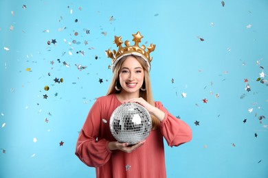 Happy young woman in party crown with disco ball and confetti on light blue background