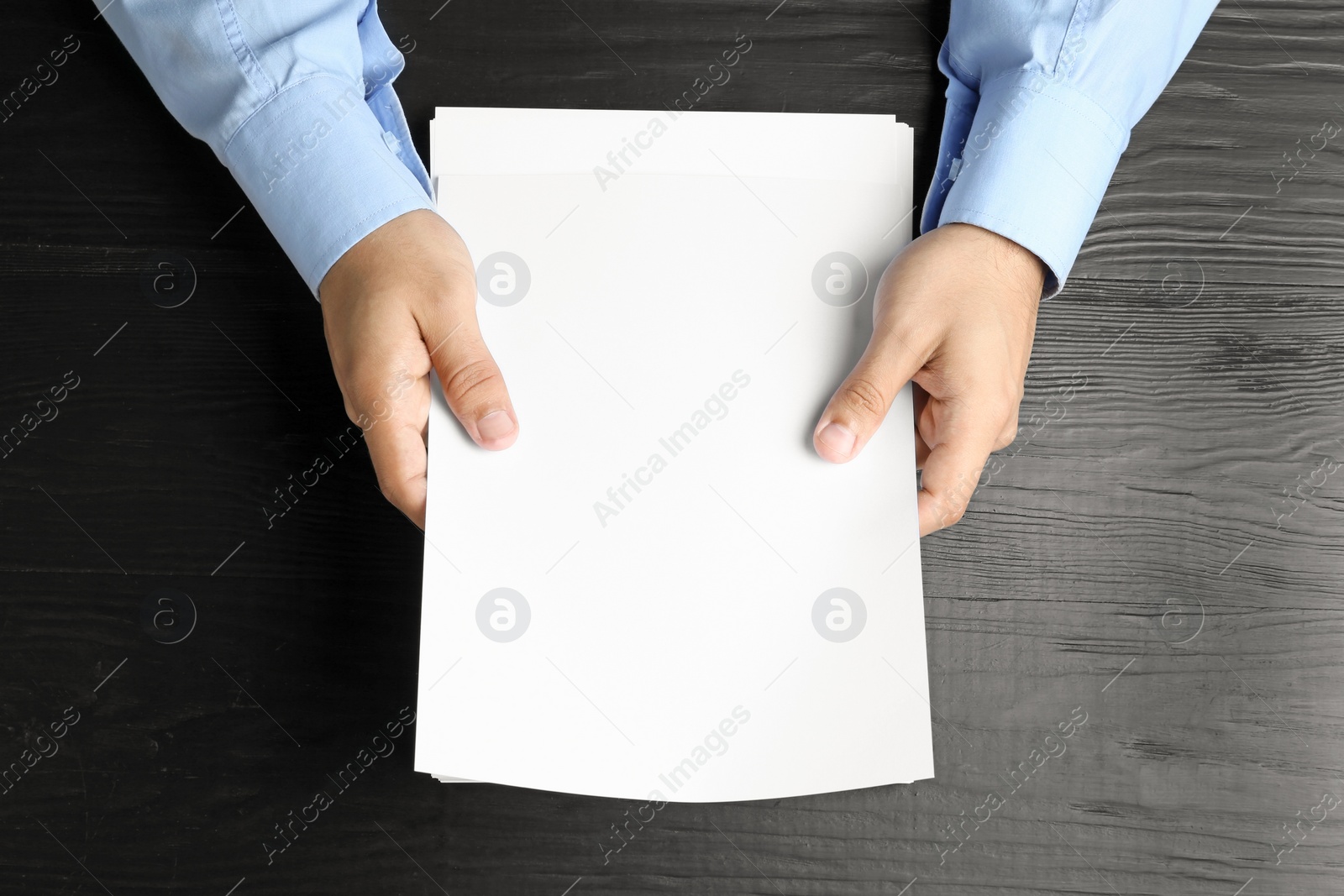 Photo of Man holding blank paper sheets for brochure at black wooden table, top view. Mock up
