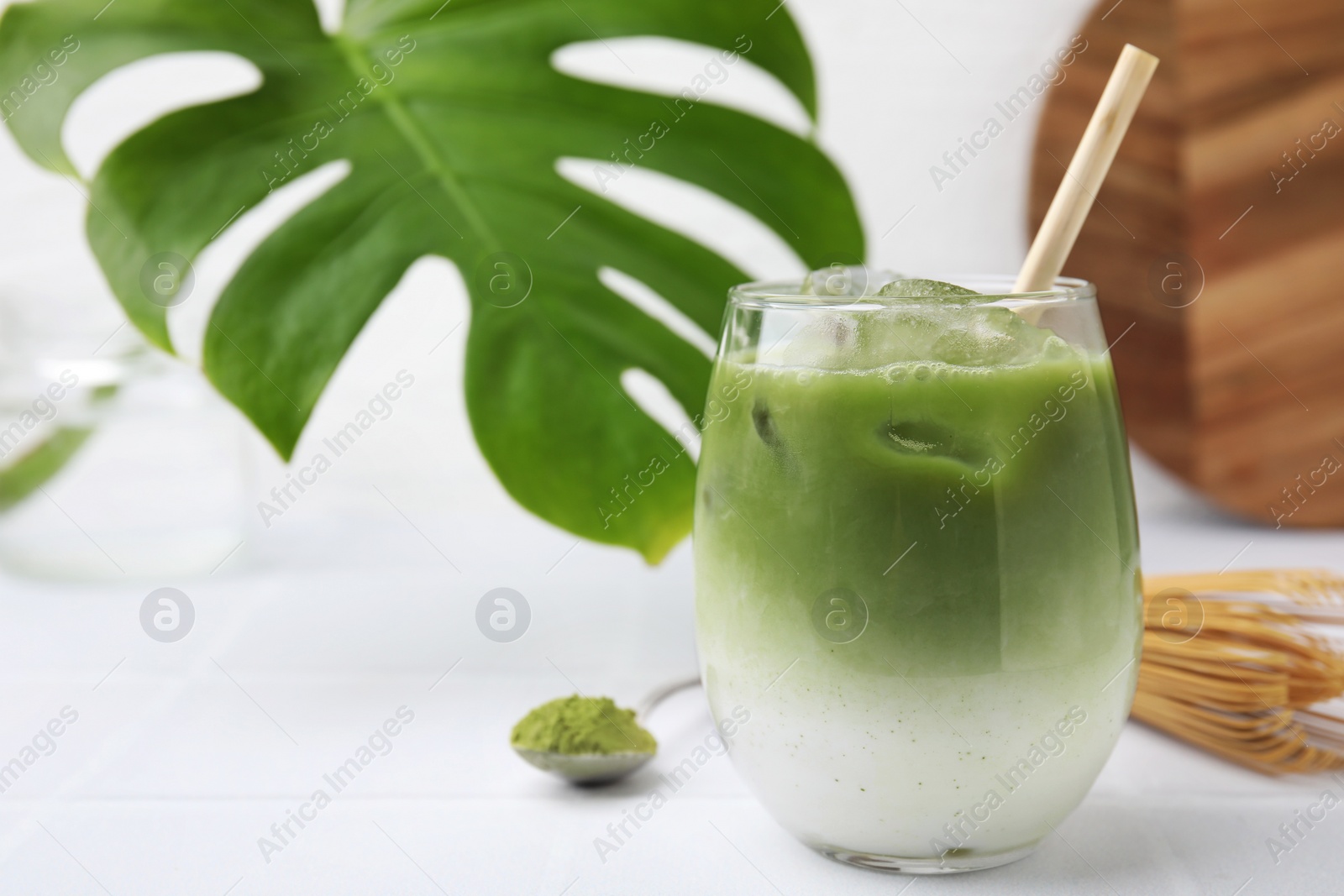 Photo of Glass of tasty iced matcha latte, leaf, bamboo whisk and powder on white tiled table, closeup. Space for text