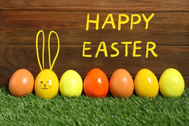 One egg with drawn face and ears as Easter bunny among others on green grass against wooden background