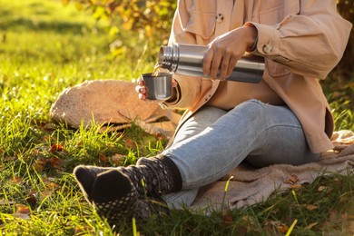 Photo of Picnic time. Woman pouring tea from thermos into cup lid on green grass outdoors, closeup