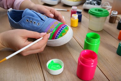 Photo of Woman painting on sneaker at wooden table, closeup. Customized shoes