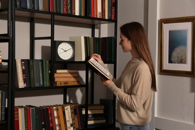 Young woman reading book near shelves in home library