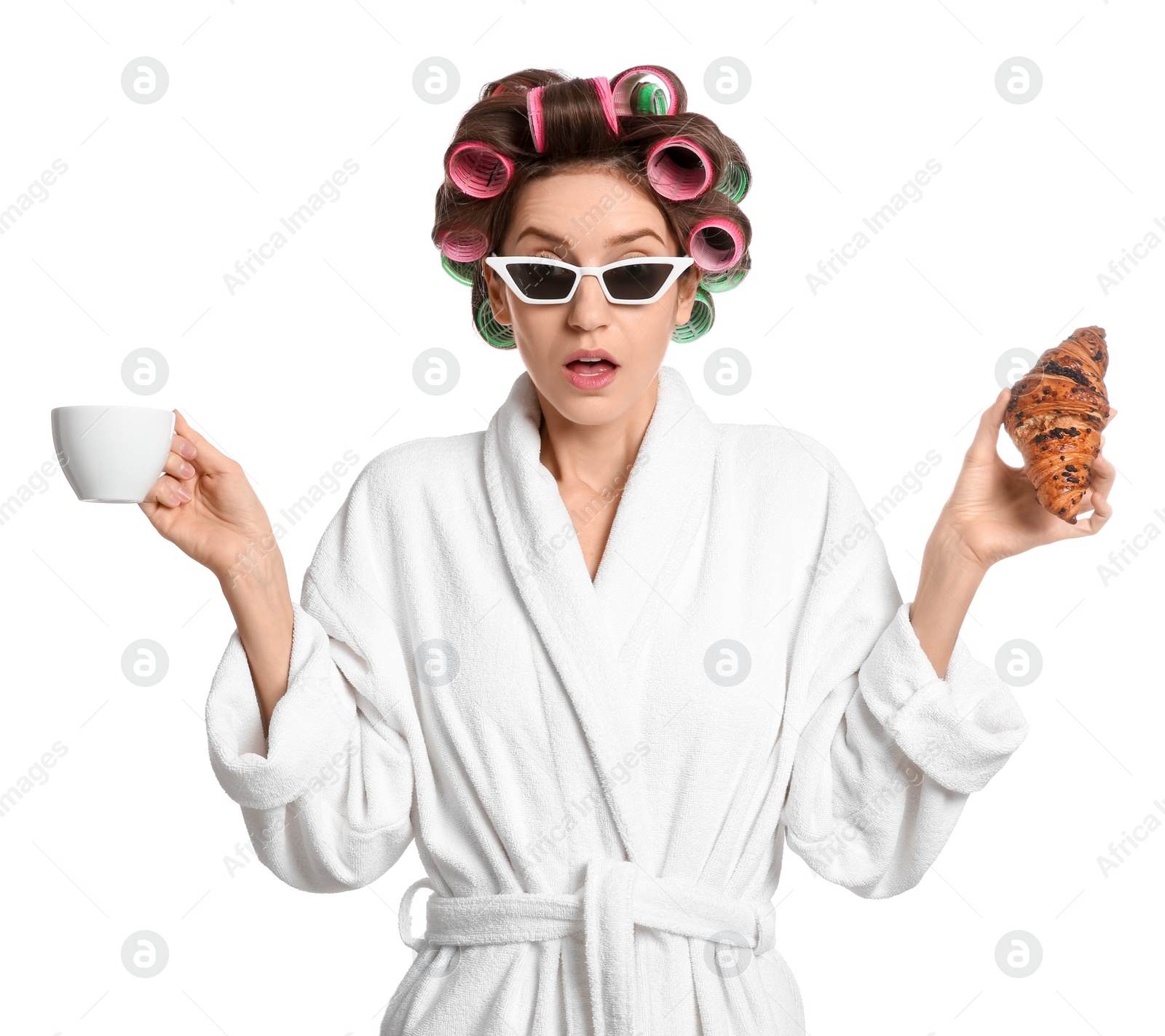 Photo of Emotional young woman in bathrobe with hair curlers holding croissant and cup of drink on white background