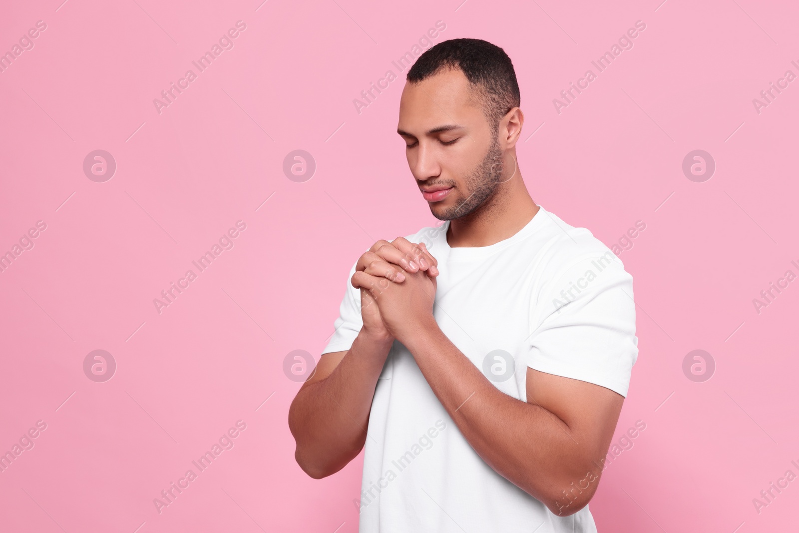 Photo of African American man with clasped hands praying to God on pink background. Space for text