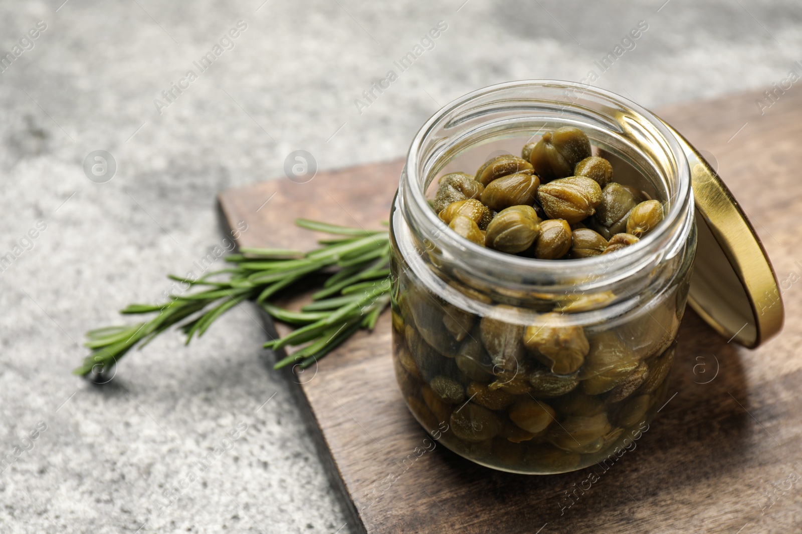 Photo of Tasty capers in glass jar and rosemary on grey table, space for text