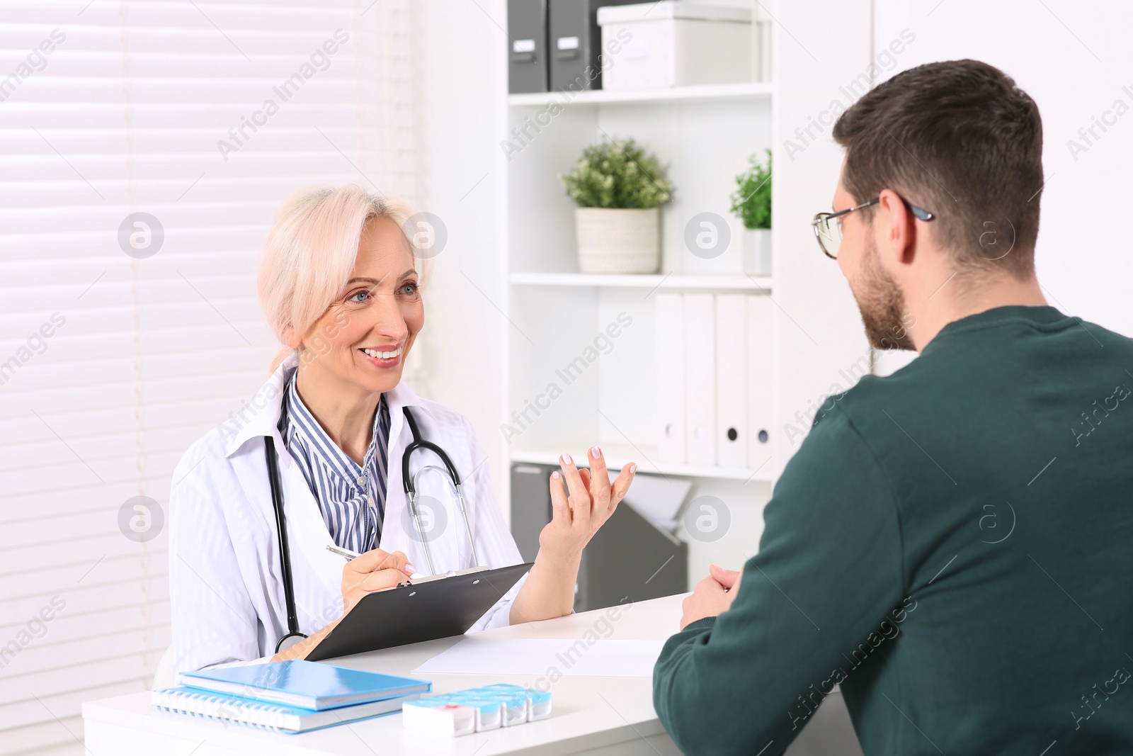 Photo of Doctor with pen and clipboard consulting patient at table in clinic