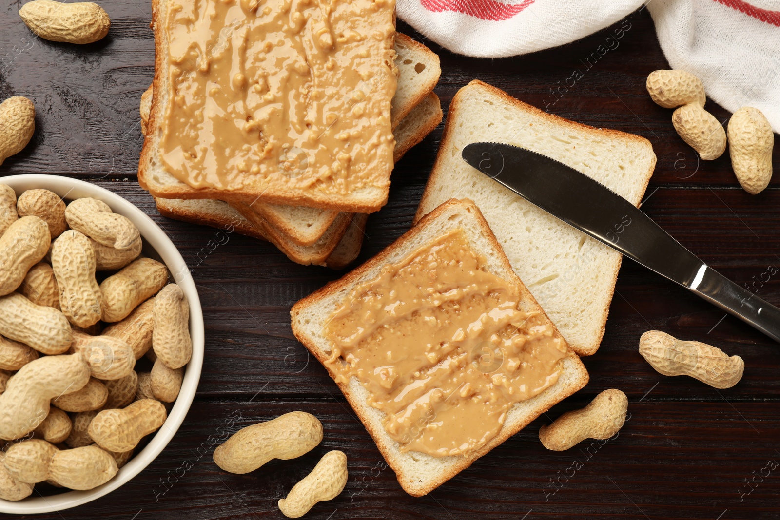 Photo of Delicious toasts with peanut butter, nuts and knife on dark wooden table, flat lay