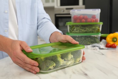 Photo of Man holding glass container with fresh broccoli at white marble table in kitchen, closeup. Food storage