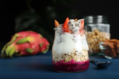 Photo of Glass jar of granola with pitahaya, yogurt and strawberries on blue wooden table, space for text