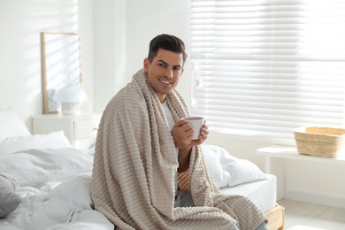 Photo of Man covered with warm beige plaid enjoying hot morning drink on bed indoors