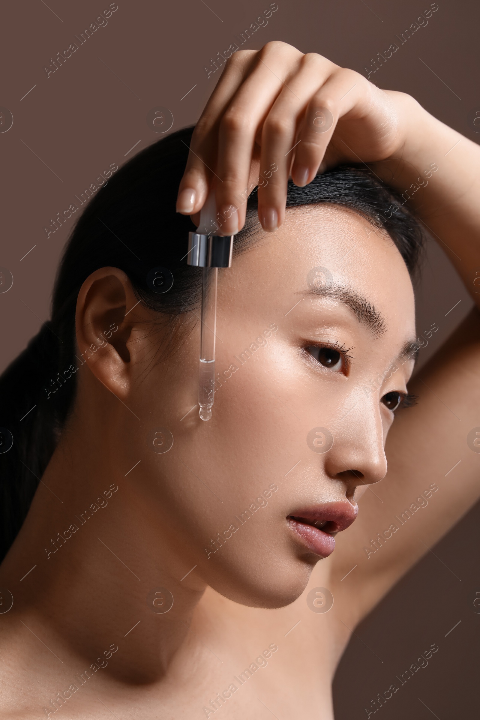 Photo of Beautiful young woman applying cosmetic serum onto her face on brown background