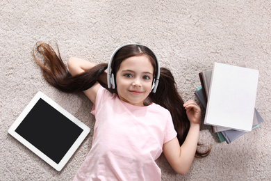 Photo of Cute little girl with headphones and tablet listening to audiobook on floor indoors, flat lay