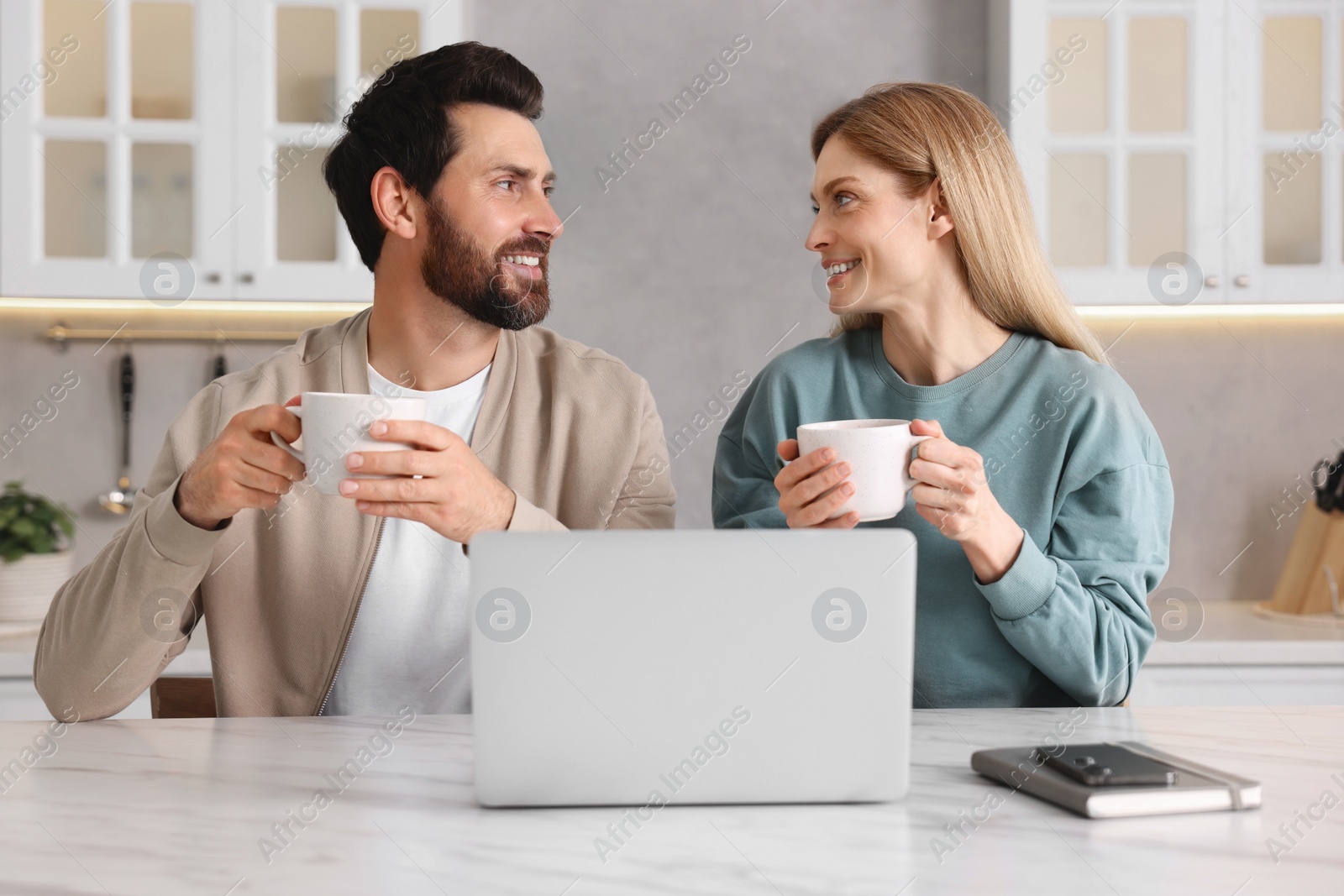 Photo of Happy couple with laptop and cups of drink at white table in kitchen