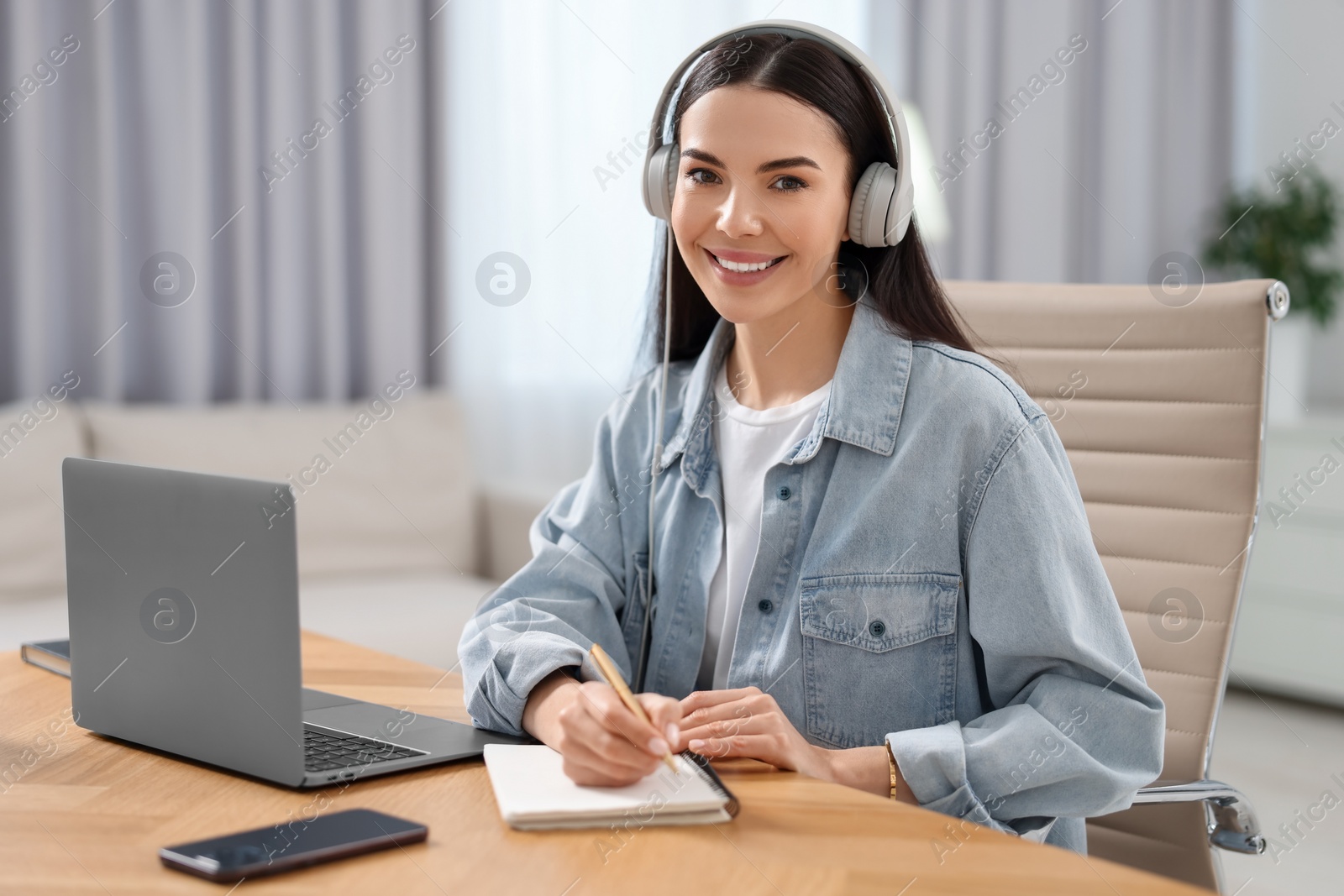 Photo of Young woman in headphones watching webinar at table in room