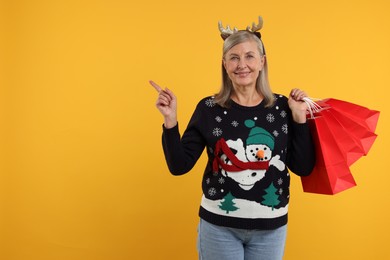 Happy senior woman in Christmas sweater and deer headband with shopping bags on orange background. Space for text