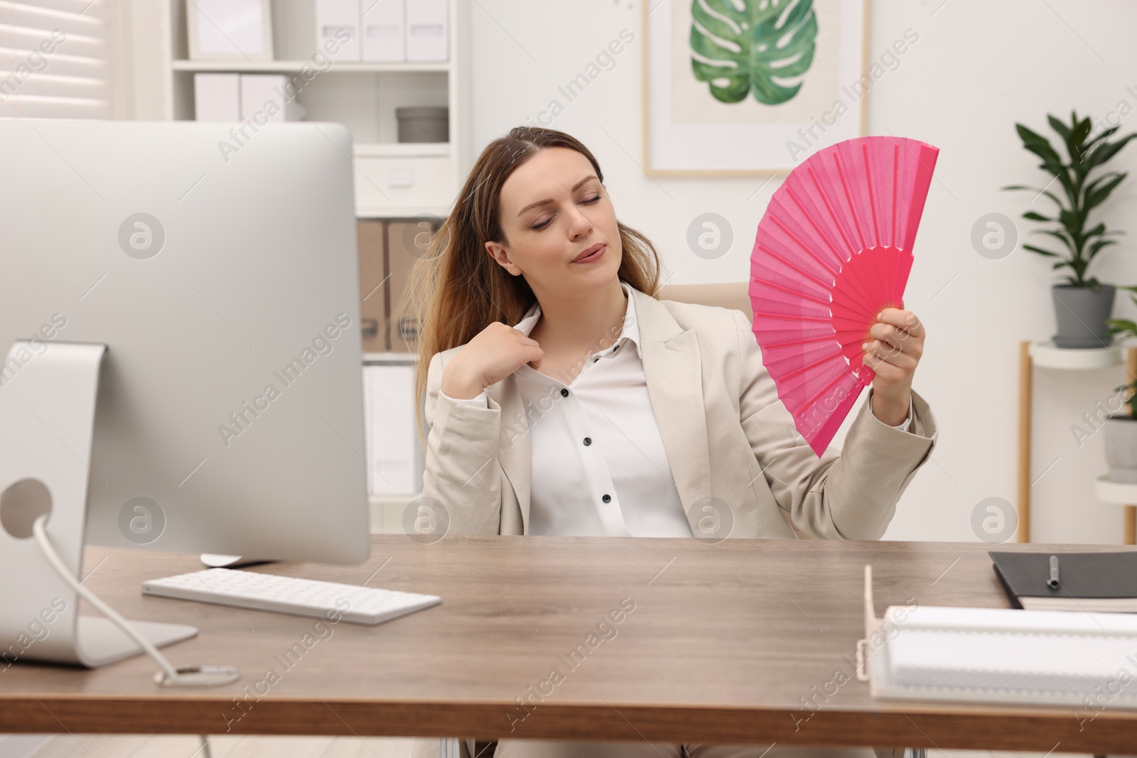 Photo of Businesswoman waving pink hand fan to cool herself at table in office