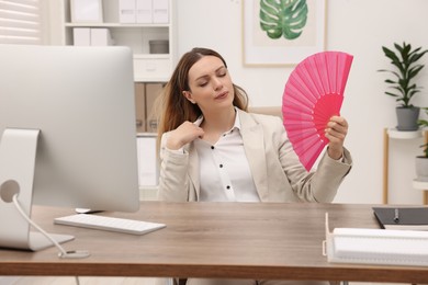 Businesswoman waving pink hand fan to cool herself at table in office