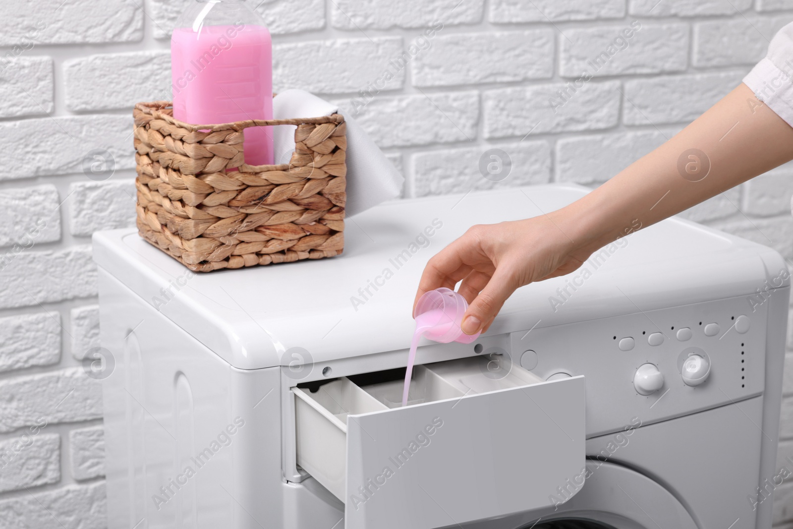 Photo of Woman pouring fabric softener from cap into washing machine near white brick wall, closeup