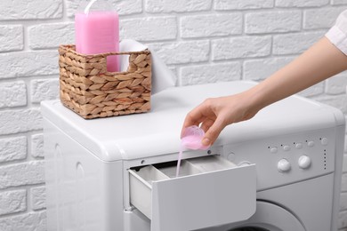 Photo of Woman pouring fabric softener from cap into washing machine near white brick wall, closeup