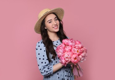 Beautiful young woman in straw hat with bouquet of peonies on pink background
