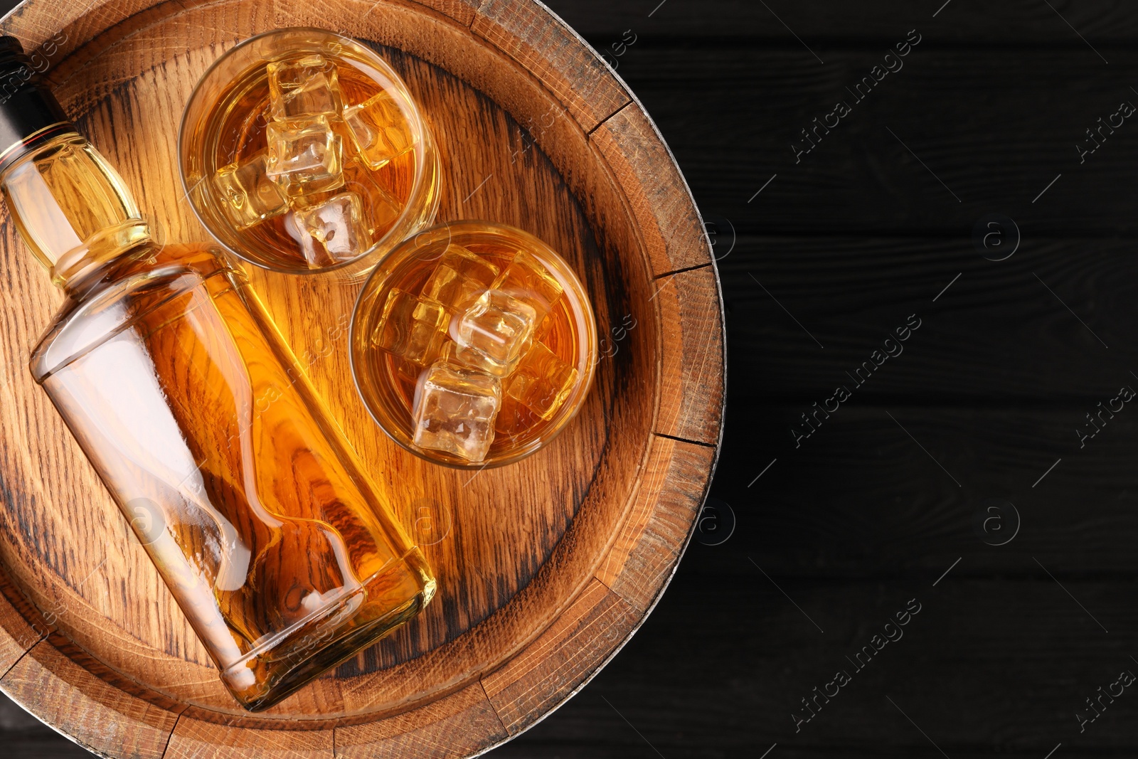 Photo of Whiskey with ice cubes in glasses, bottle and barrel on black wooden table, top view. Space for text