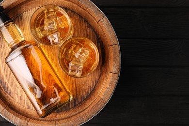 Whiskey with ice cubes in glasses, bottle and barrel on black wooden table, top view. Space for text