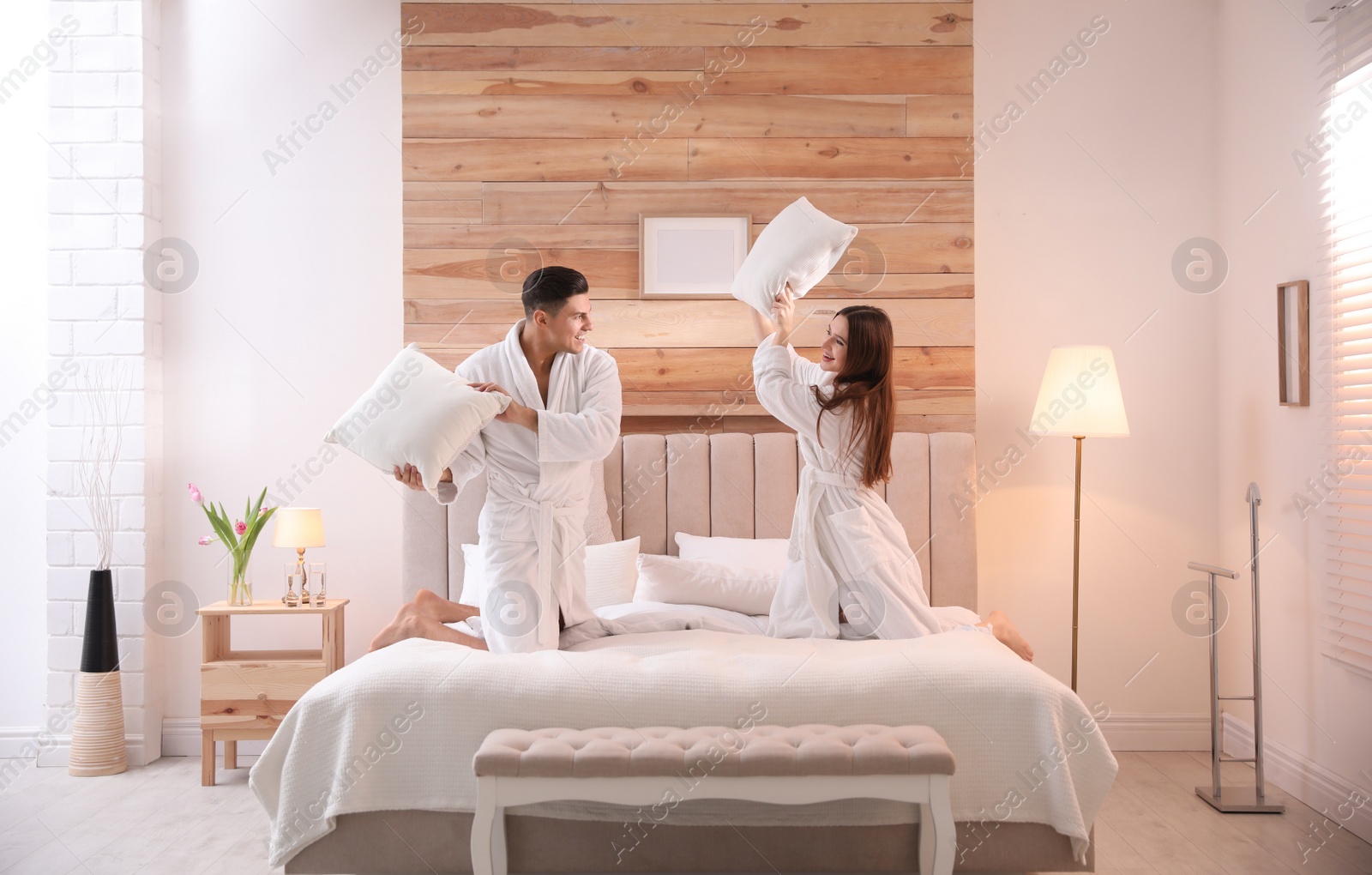 Photo of Happy couple in bathrobes having pillow fight on bed at home