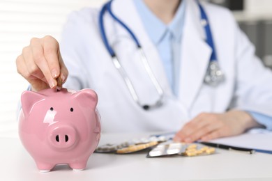 Doctor putting coin into piggy bank at white table indoors, closeup