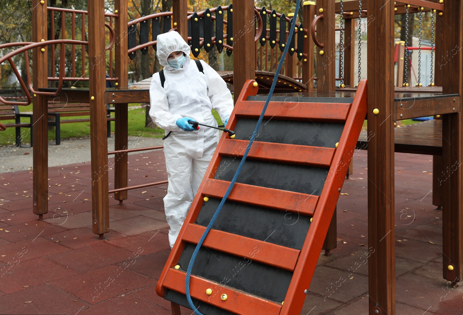 Photo of Woman wearing chemical protective suit with disinfectant sprayer on playground. Preventive measure during coronavirus pandemic