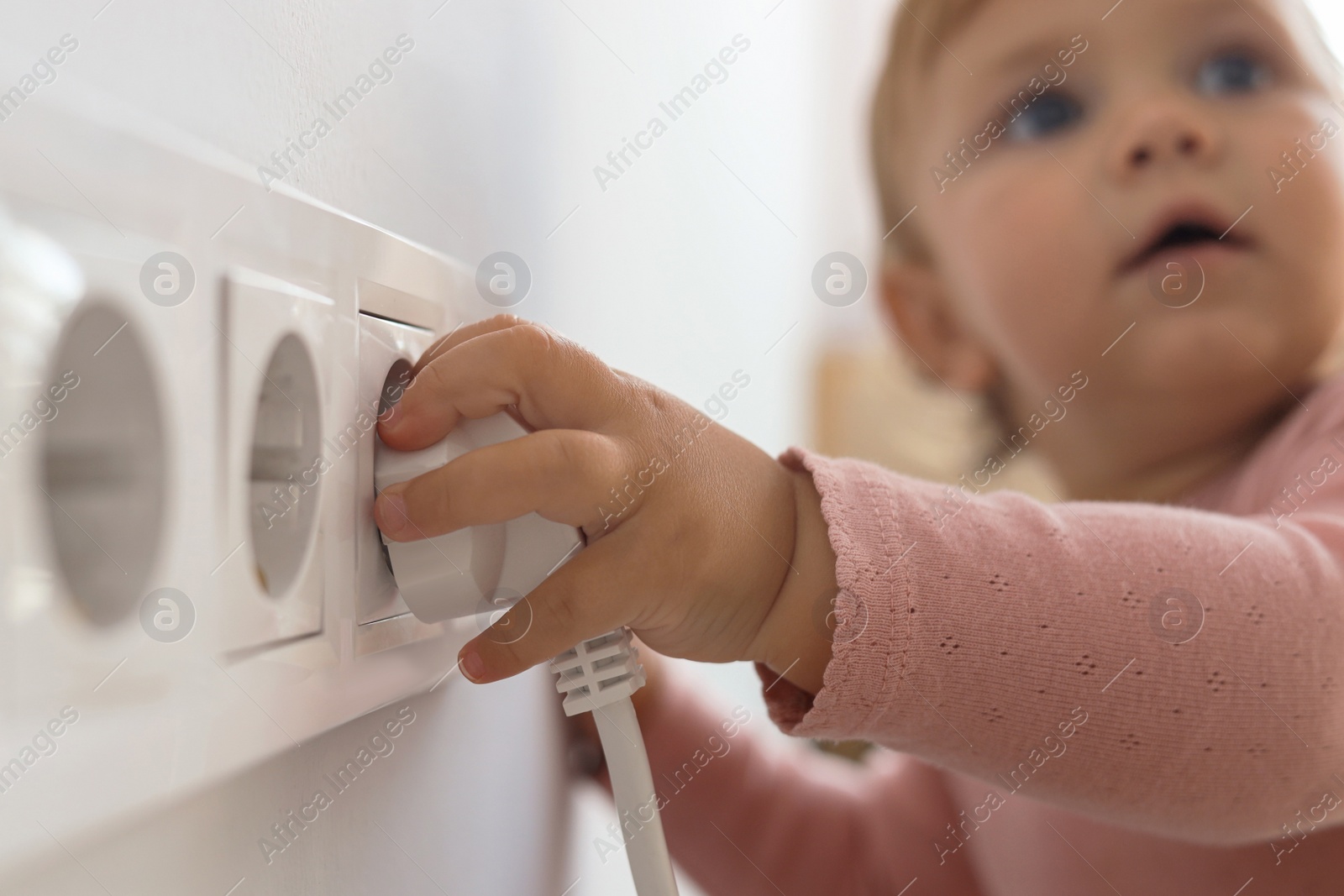 Photo of Cute baby playing with electrical socket and plug at home, focus on hand. Dangerous situation