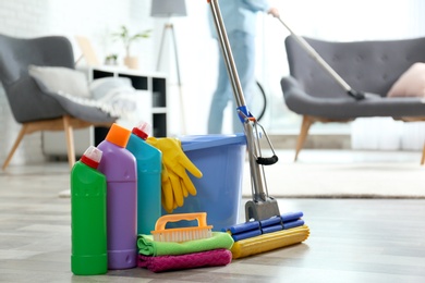 Photo of Detergents, bucket and mop on floor with janitor vacuuming carpet indoors