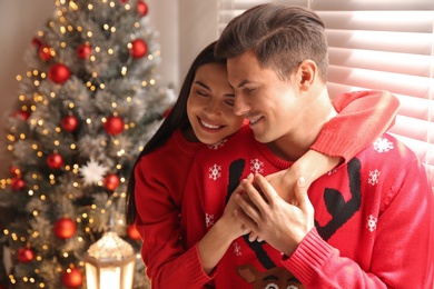 Photo of Happy couple in warm sweaters near Christmas tree indoors