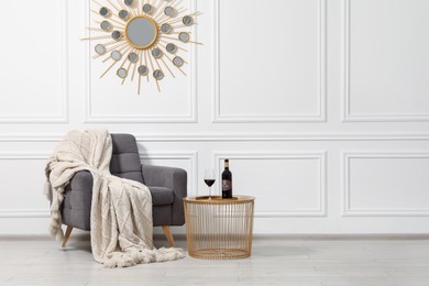 Photo of Comfortable armchair, blanket and wine on side table near white wall, space for text. Interior design