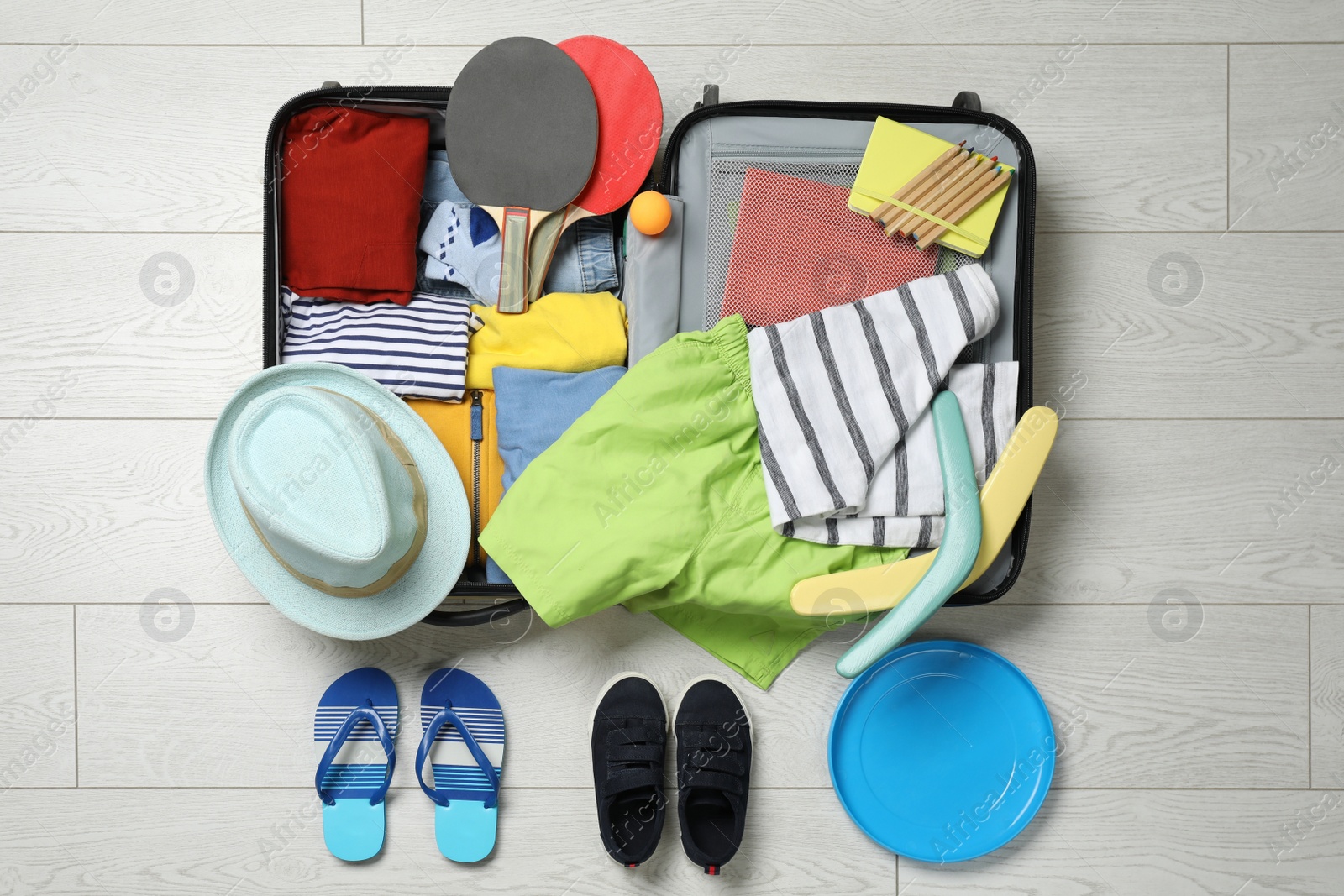 Photo of Open suitcase with stylish clothes, accessories, sport equipment and shoes on floor, flat lay. Summer vacation