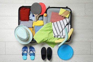 Open suitcase with stylish clothes, accessories, sport equipment and shoes on floor, flat lay. Summer vacation