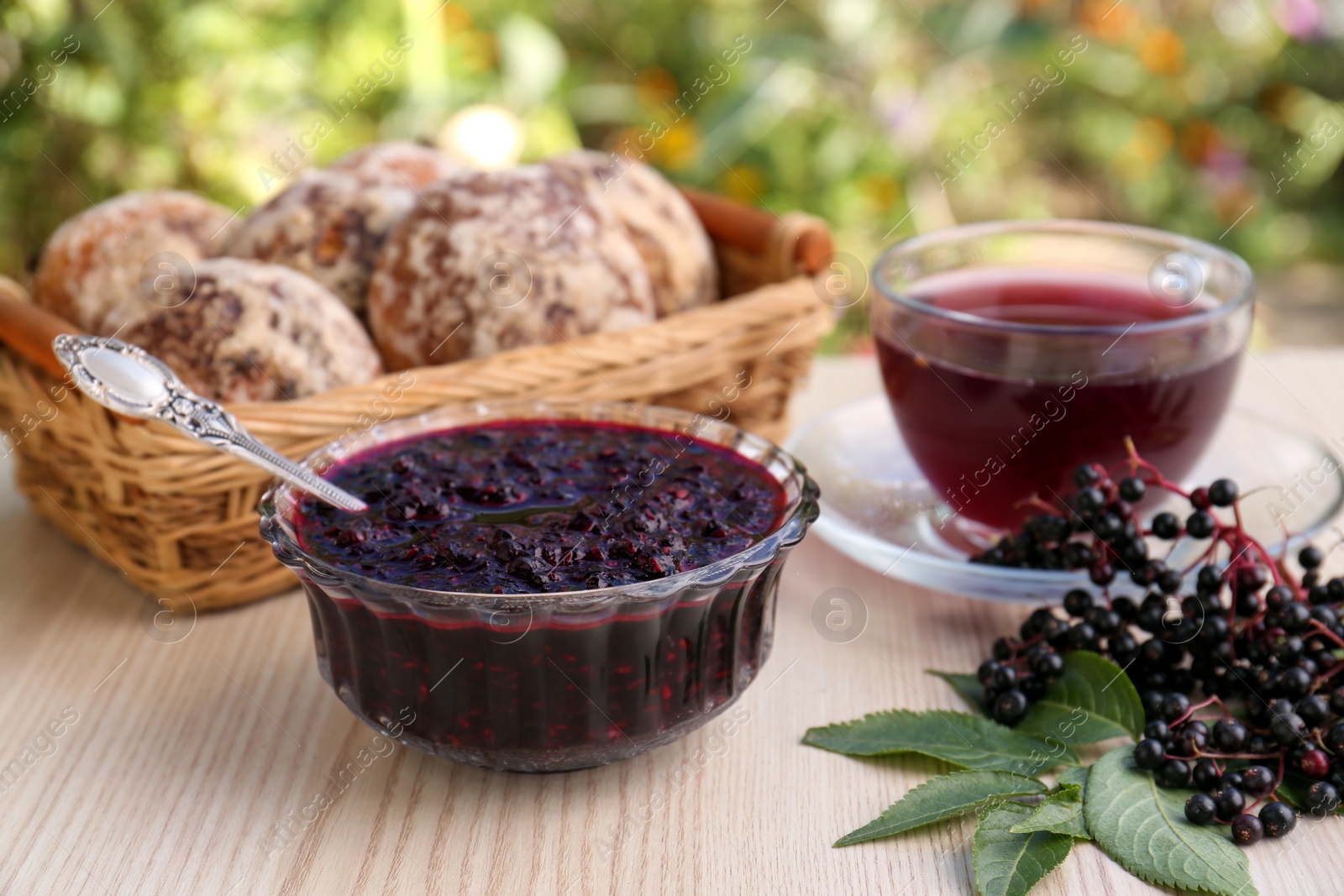 Photo of Elderberry jam, glass cup of tea, tasty cookies and Sambucus berries on wooden table outdoors